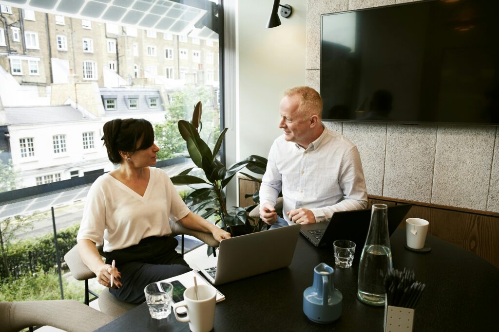 Home Two business professionals engaging in a team meeting in a modern London office with a large window view.